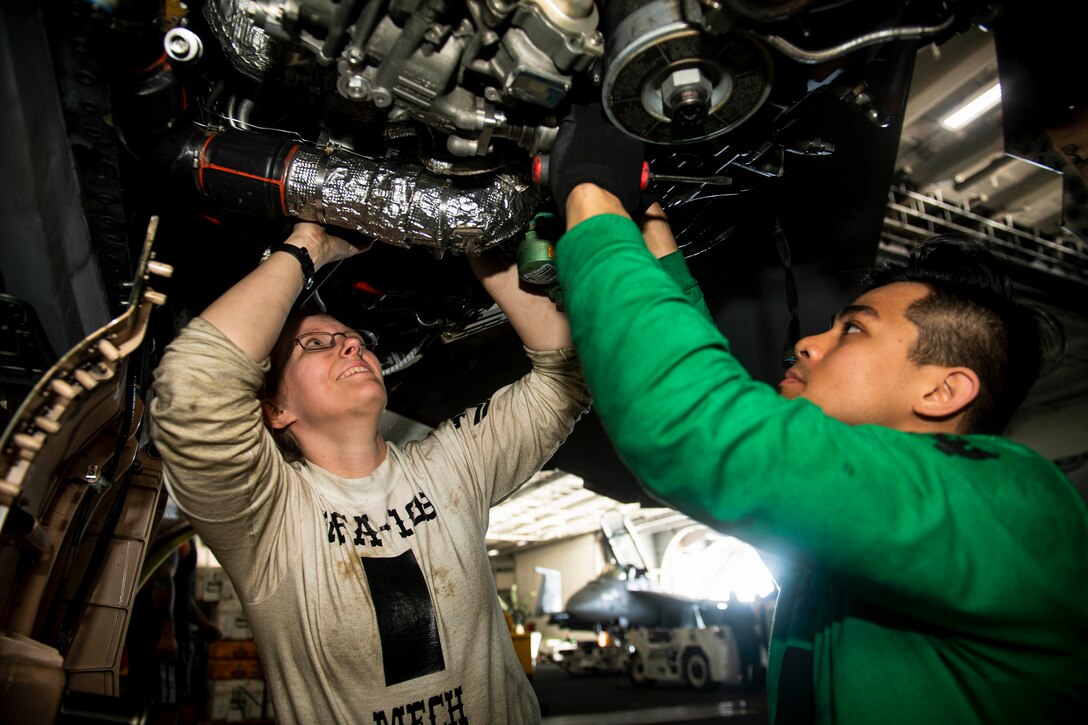 Two sailors work underneath an aircraft.