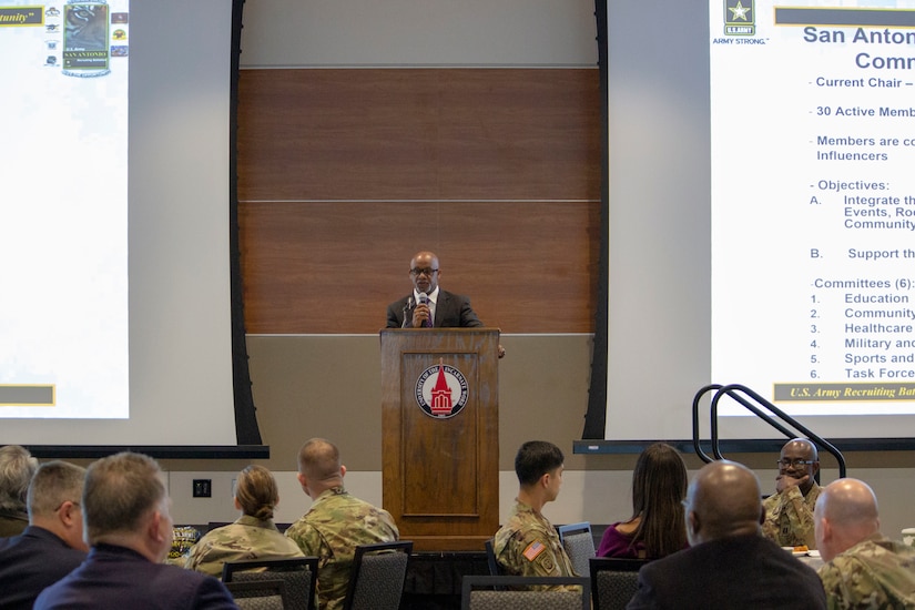 Black male in dark suit, white suit and red tie stands at wooden podium on stage with men and women sitting in crowd in suits and green camouflage uniforms. Two large screens hang to each side of photo.