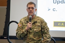 White male in green camouflage uniform holds microphone stands in front of large screen.