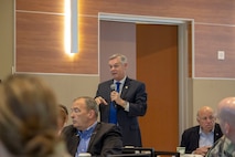 White man in dark suit and blue tie stands with microphone in room with people sitting.