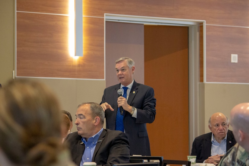 White man in dark suit and blue tie stands with microphone in room with people sitting.