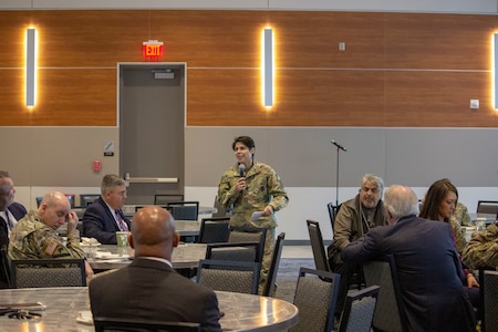 White female in green camouflage uniform holding a microphone stands in the middle of a room filled with tables and men and women both in suits and green camouflage uniforms.