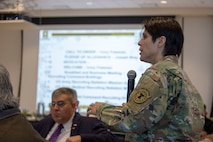 White female in green camouflage stands holding a microphone, a white man in a suit sits in the audience with a large screen and slide up in the background.
