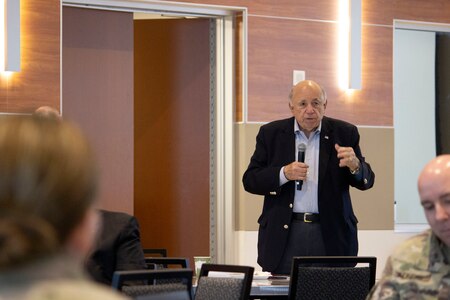 White male in dark suit and white shirt stands holding a microphone two individuals in green camouflage sit in the wood paneled conference room.