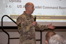 Bald male in green camouflage uniform stands holding michrophone in front of a screen.