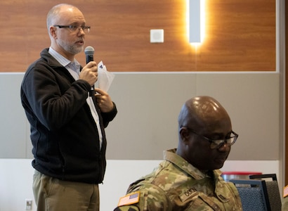 White male with beard in white shirt, black jacket and kaki pants holds microphone and papers, a black man with glasses in green camouflage uniform sits in front of him.