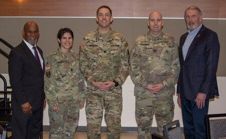 Two men in dark suits stand with two men and one woman in green camouflage uniforms.