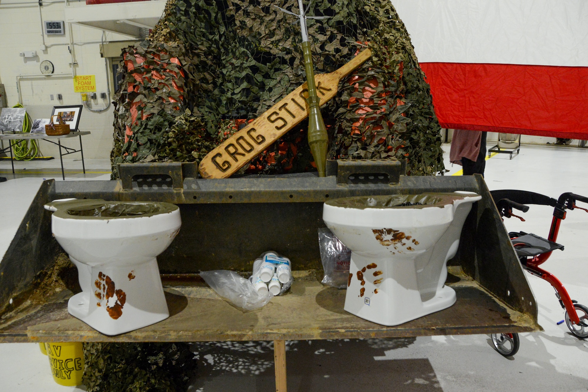 Two sanitary toilet bowls sit on military construction equipment. An inert missile and a paddle labeled Grog Stick lean against camouflage netting.