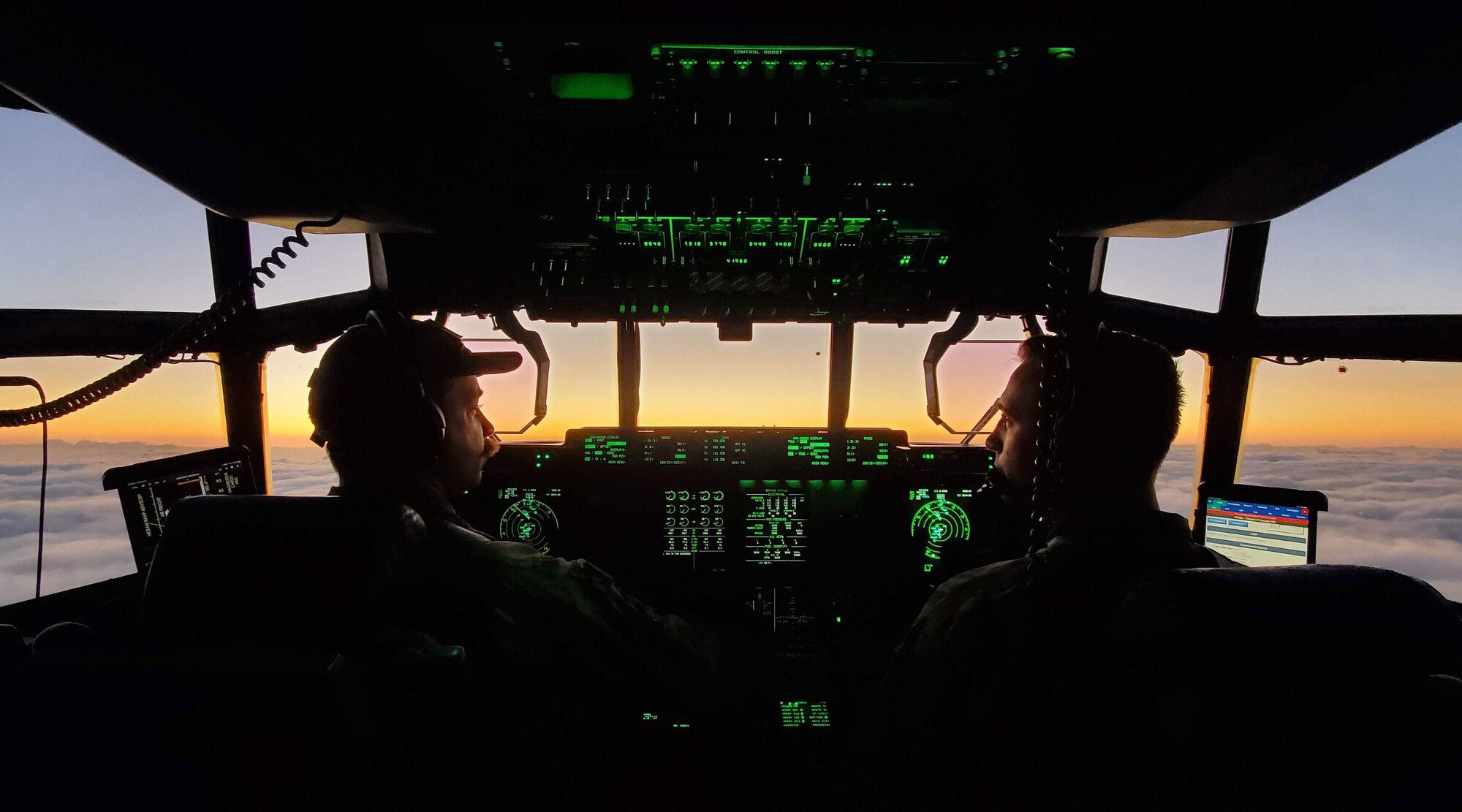 Two Airmen fly a plane above the clouds with a sunset in the background.