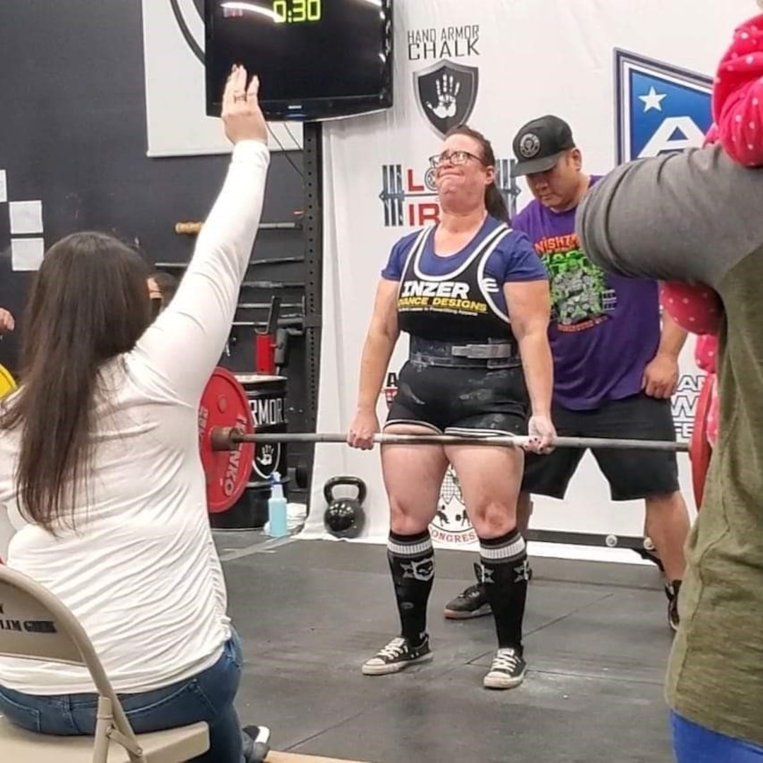 Lady in audience raises her hand while a female powerlifter deadlifts.