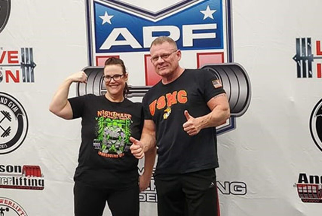 Man and woman stand in front of powerlifting photo banner