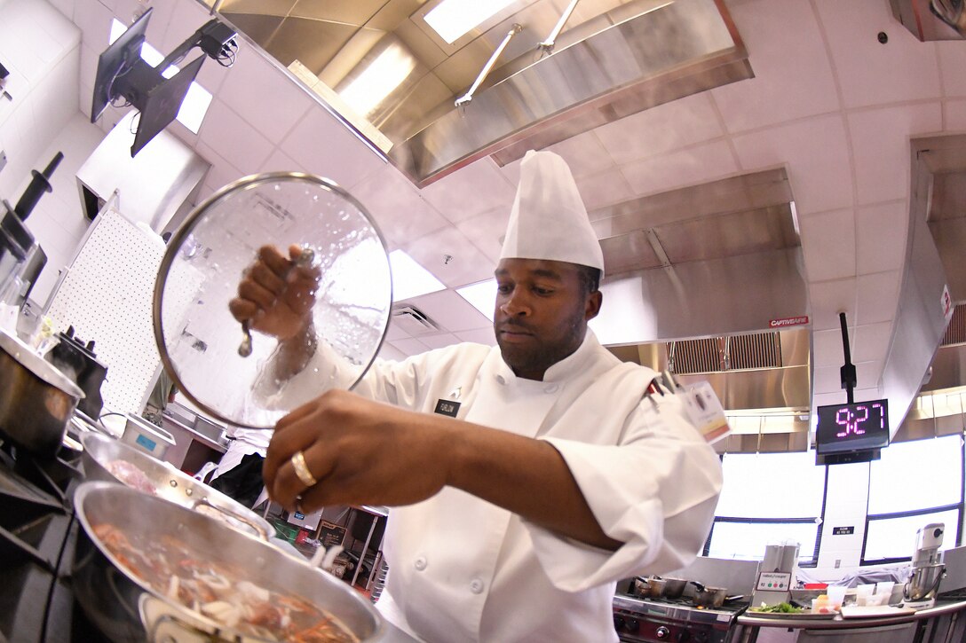 A man in a chef's uniform adds some spices to a pot.