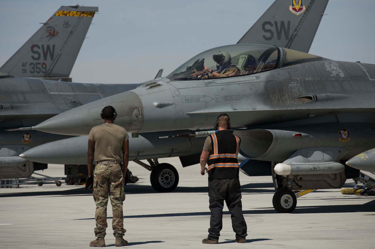 Two service members stand on a flight line near a military combat aircraft.