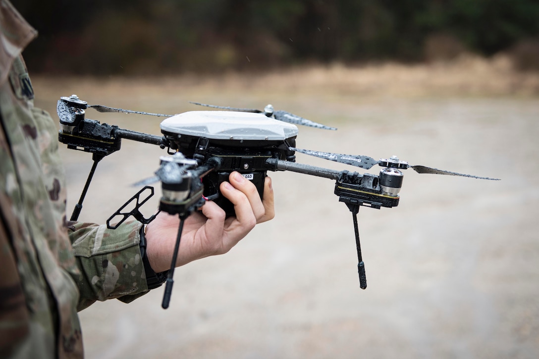 A soldier holds a drone on flat dirt terrain.