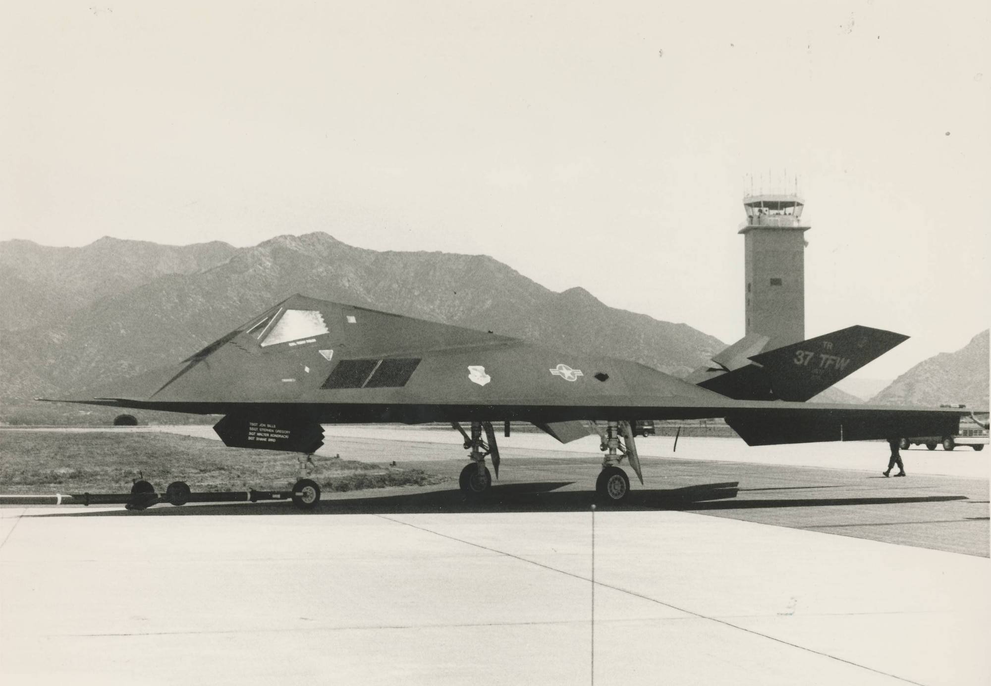 An F-117 on the Hill AFB flight line. The base's air traffic control tower and the Wasatch Mountains appear in the background.