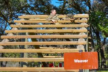 Male in green camouflage uniform hangs between two wood beams on an outdoor obstacle that has tiered wood beams.