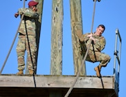 Black male in red beret and green camouflage uniform stands holding rope while white male in green camouflage uniform grabs onto the rope on an outdoor wood obstacle.