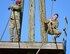 Black male in red beret and green camouflage uniform stands holding rope while white male in green camouflage uniform grabs onto the rope on an outdoor wood obstacle.