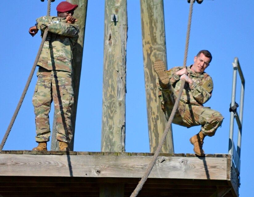 Black male in red beret and green camouflage uniform stands holding rope while white male in green camouflage uniform grabs onto the rope on an outdoor wood obstacle.