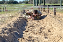 Brown-haired male in green camouflage uniform crawls in dirt under barbed wire.