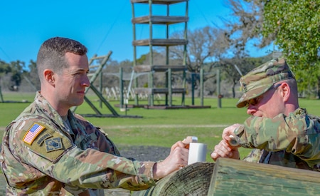 One male in green camouflage uniform and no hat stands with another male in green camouflage at an outdoor wood obstacle.