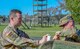 One male in green camouflage uniform and no hat stands with another male in green camouflage at an outdoor wood obstacle.