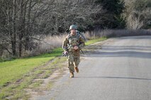 Male in green camouflage uniform and gray helmet runs down a dirt road.