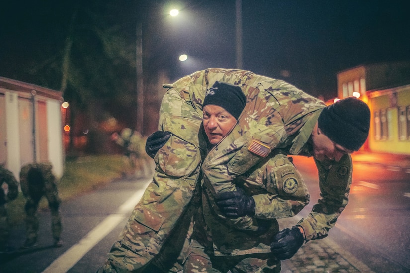 A soldier dressed in fatigues carries another soldier over his shoulders.