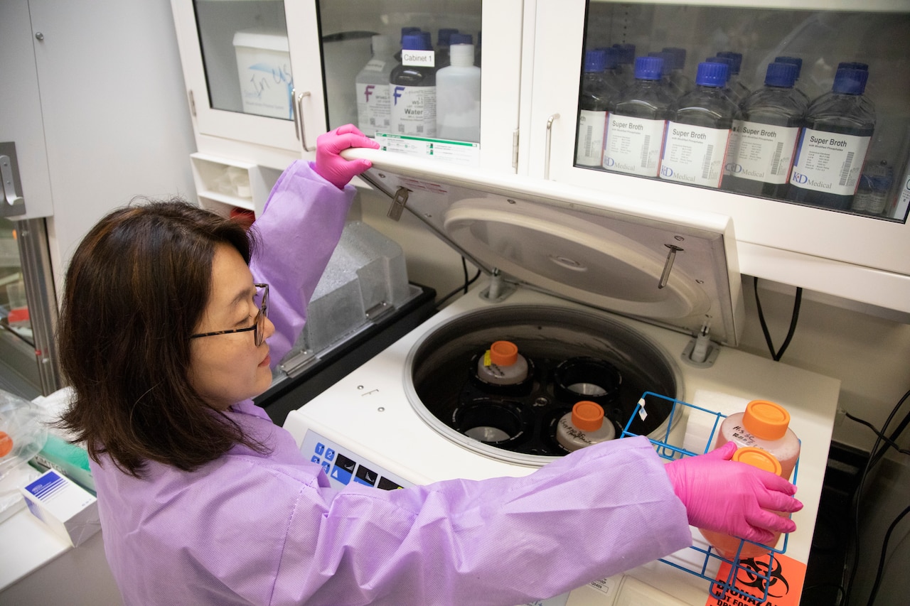 A woman in a laboratory places jars with orange lids into a centrifuge.