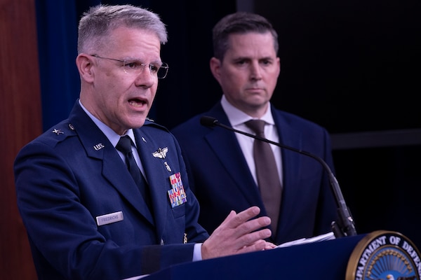 An Air Force officer speaks from behind a podium. A civilian man stands to his left.
