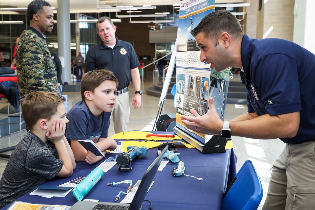 Cartridge Actuated Device/Propellant Actuated Device (CAD/PAD) Fleet Support Team/Mishap Investigation Support Team lead Nick Schombs discusses CAD/PAD technology with Mason Whetzel, 5,(bottom left) and Dylan Whetzel, 8, during the annual HITS Expo at St. Charles High School, Saturday. (Photo by Matt Poynor)