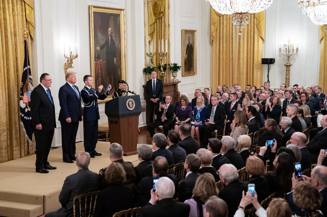 Three men stand on a stage in front of a large group of people during a ceremony.