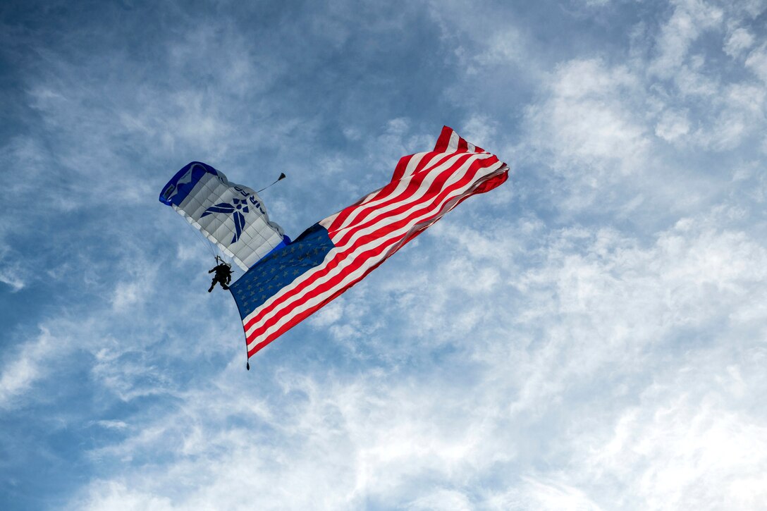 An airman with a parachute descends in blue sky while displaying an American flag