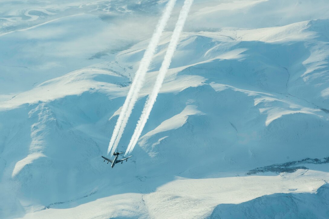 A Sentry aircraft creates white smoke trails while flying above snow-covered mountains.