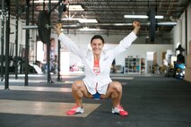 White female in white lab coat and orange shirt squats with a barbell over her head.