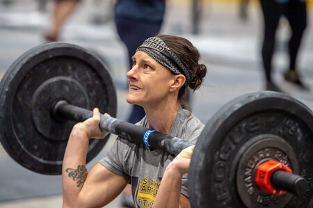 White female in gray and yellow t-shirt and black and white headbands holds barbell with eights