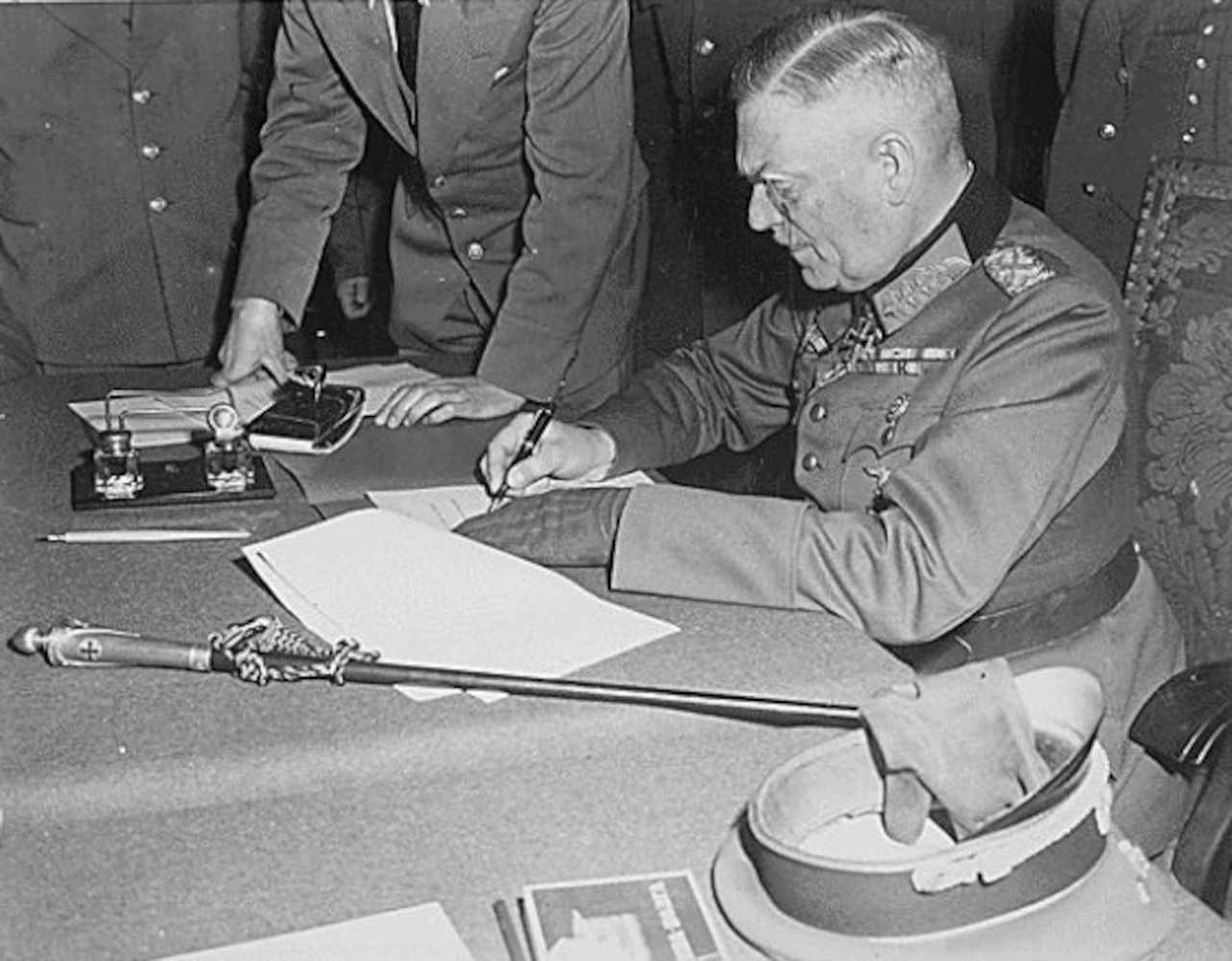 A military man signs some papers while others look on.
