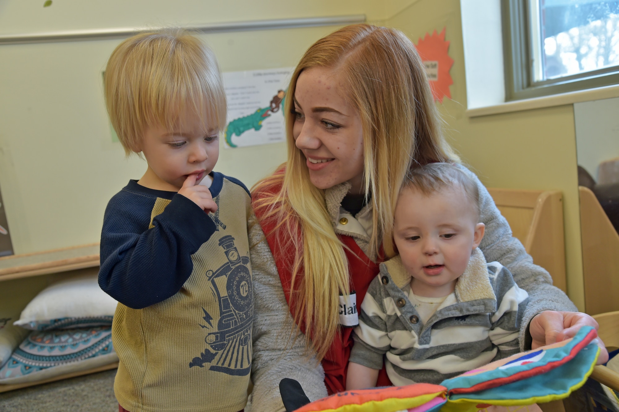 Claire Silver (center), 319th Force Support Squadron, child development program assistant, reads with toddlers at the Child Development Center on Grand Forks Air Force Base, N.D., March 10, 2020. Effective 1 April, the CDC will change its hours to open at 0700 and will close at 1730. The change is necessary to allow for mandated scheduled breaks for caregivers, curriculum planning and establishes continuity of caregivers in the classrooms. (U.S. Air Force photo by Tech. Sgt. Kamaile Casillas)