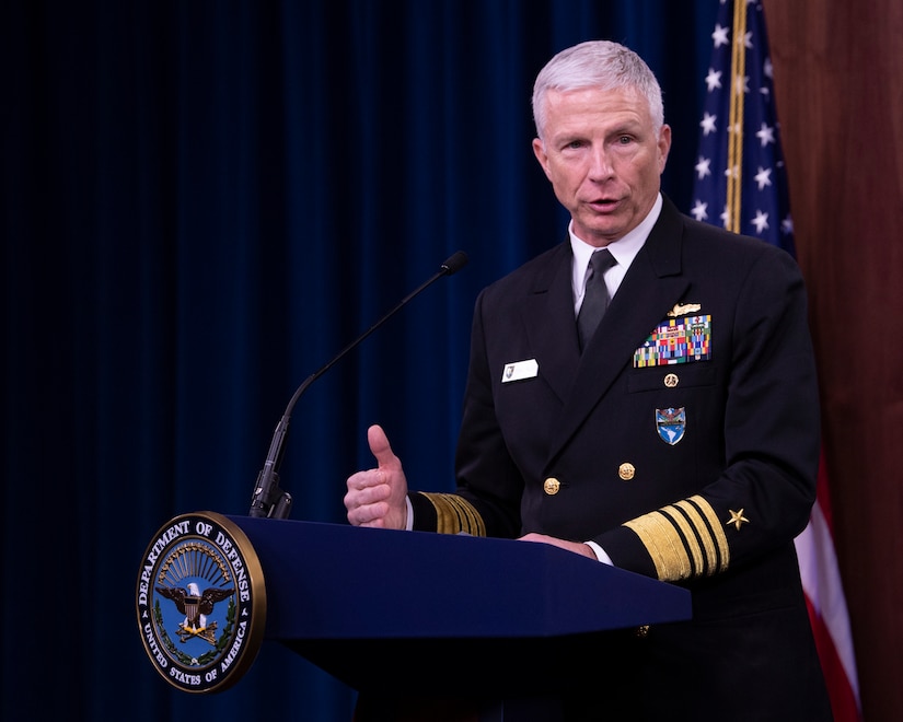 A man in a military uniform stands behind a lectern.  Behind him is an American flag.