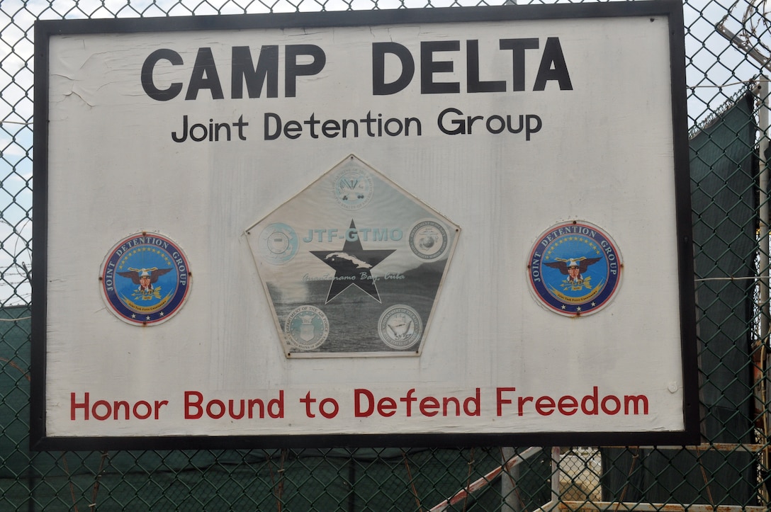 A sign hangs on a fence. It reads “Camp DELTA. Joint detention group.  Honor Bound to Defend Freedom.”