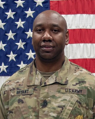 Black male in green camouflage uniform stands in front of an American Flag with the red and white stripes and the blue background with white stars.
