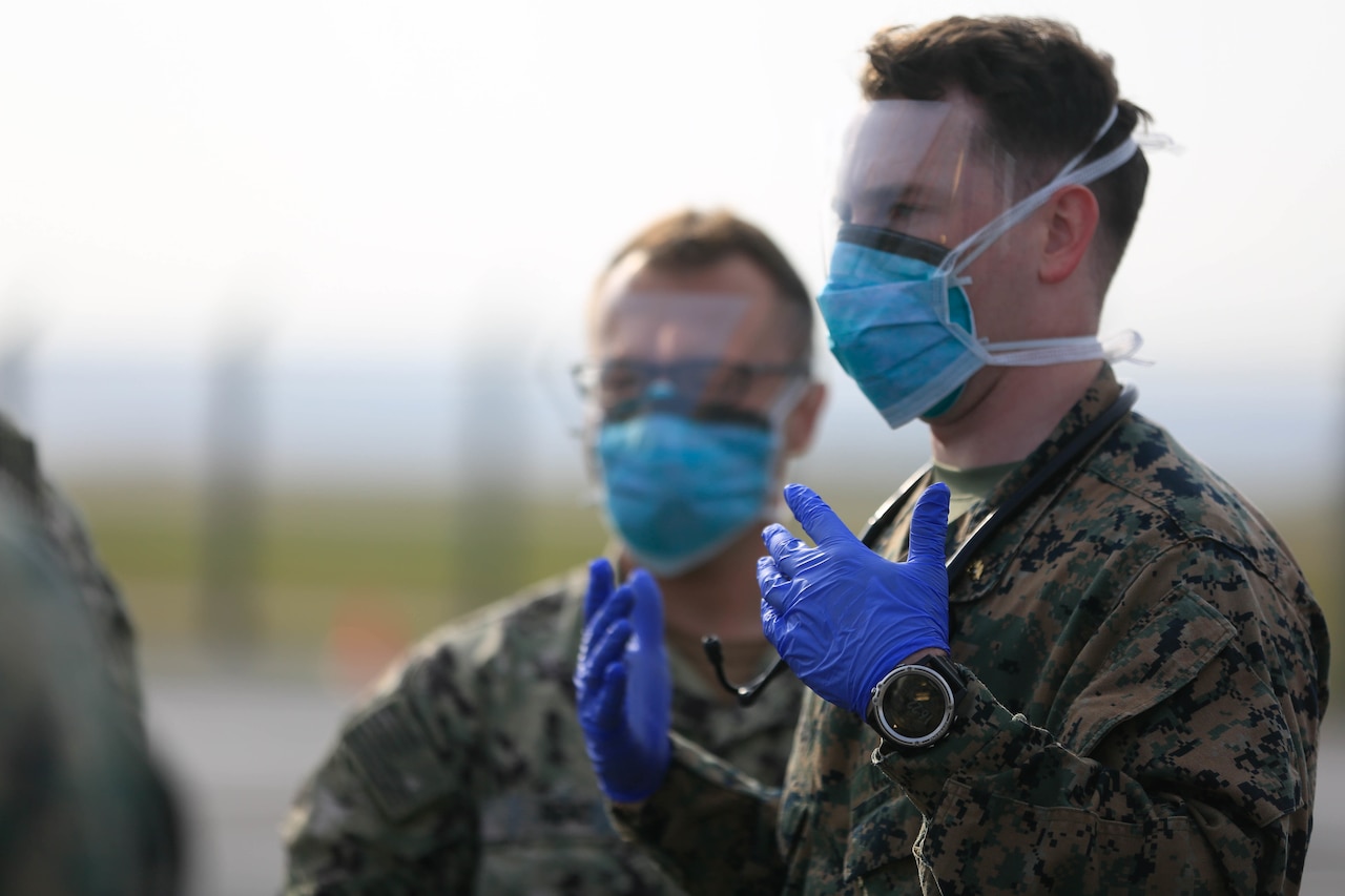 Two men wearing gloves and surgical masks over their noses and mouths look into the distance.