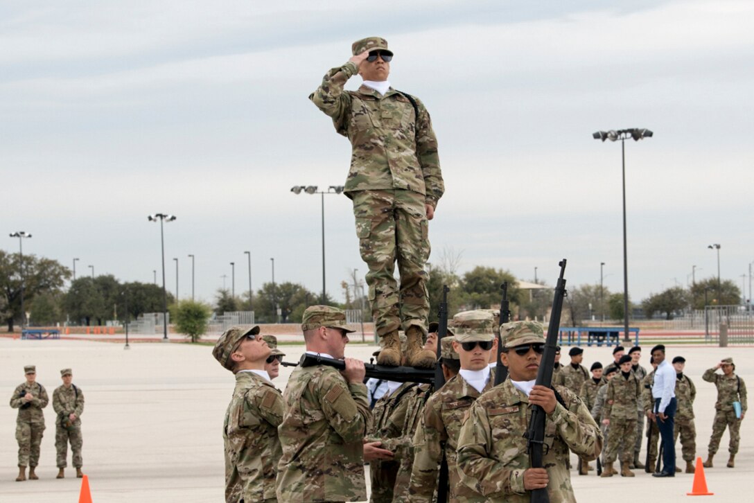An airman leans back and salutes while standing atop rifles held by a cluster of fellow airmen on a paved lot.