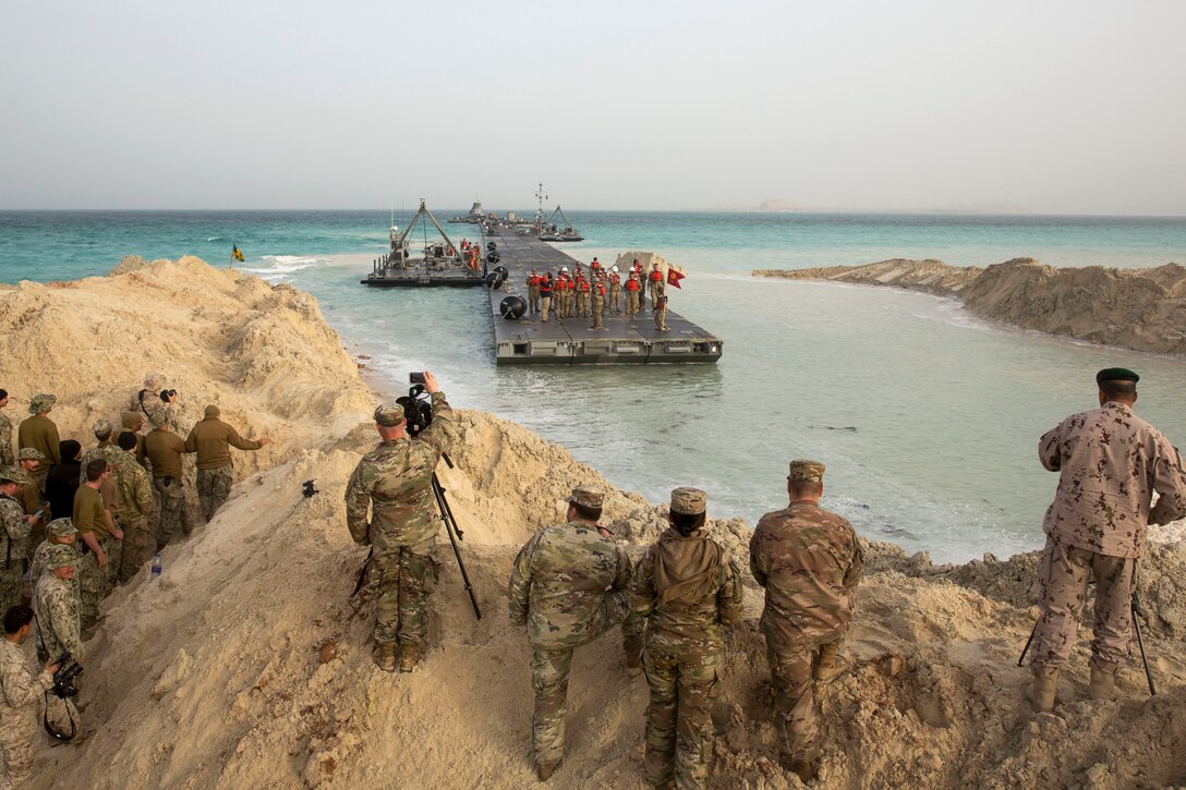 U.S. and foreign service members watch on a dirt hill as a floating causeway approaches.