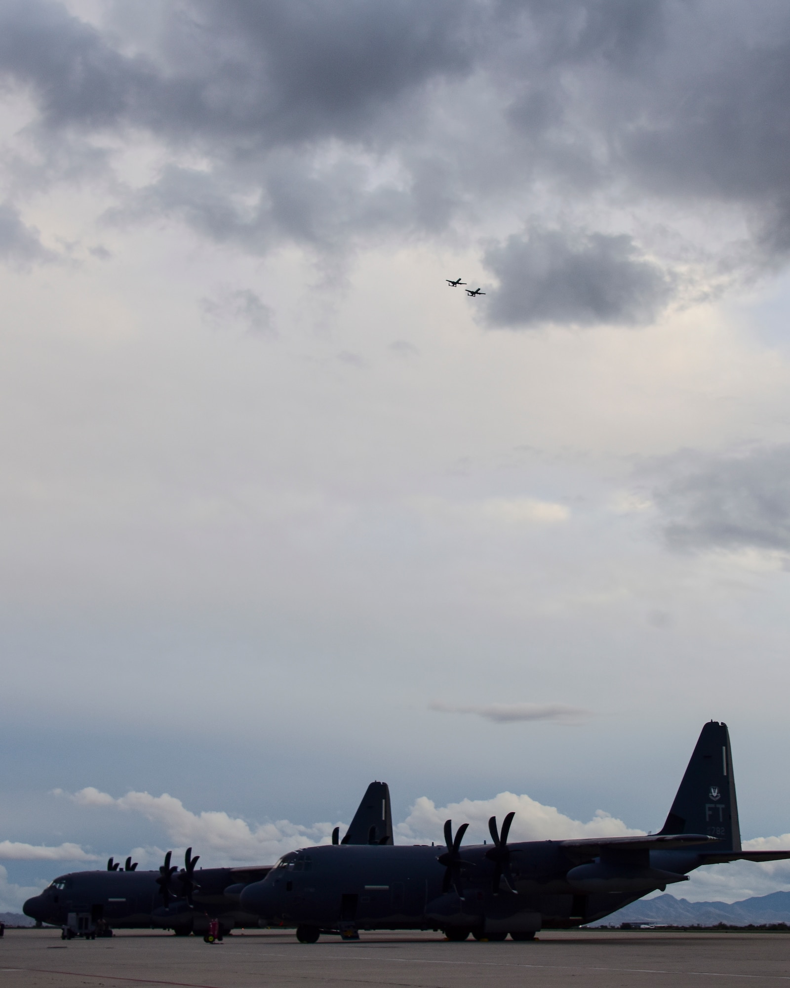 A photo of HC-130Js sitting on the flight line as two A-10s fly over