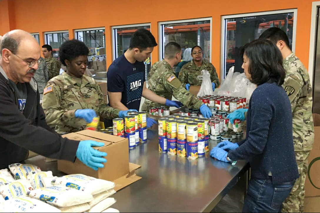 Soldiers and airmen work with people in civilian clothes to organize canned foods while standing at a metal table.