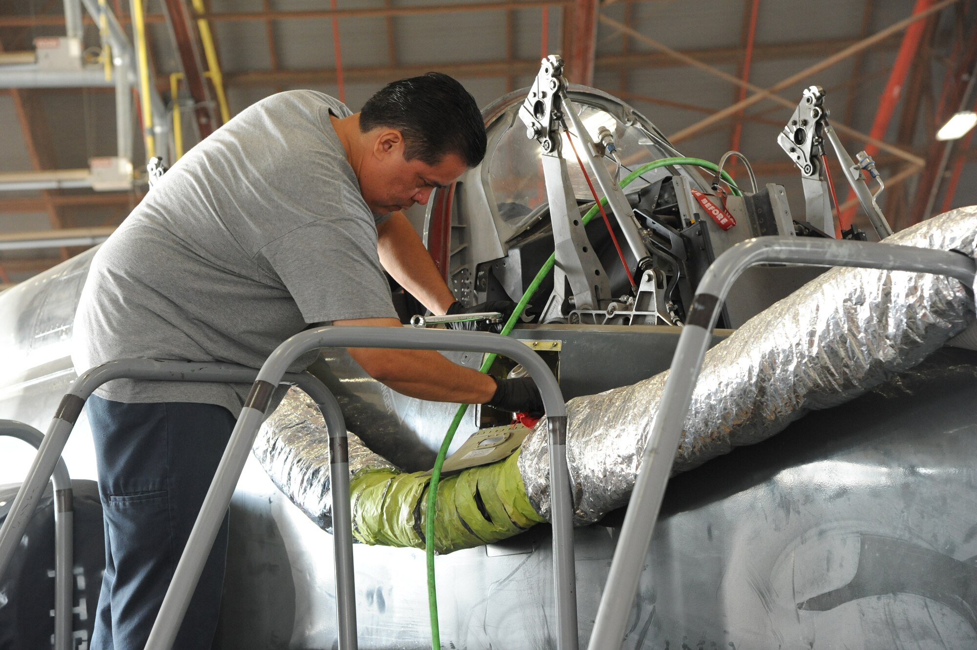 An Aircraft maintainer standing on a platform to conduct maintenance on a T-38 cockpit inside a hangar.