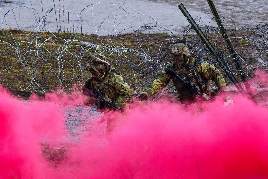 Two soldiers run toward a cloud of pink smoke near barbed wire.