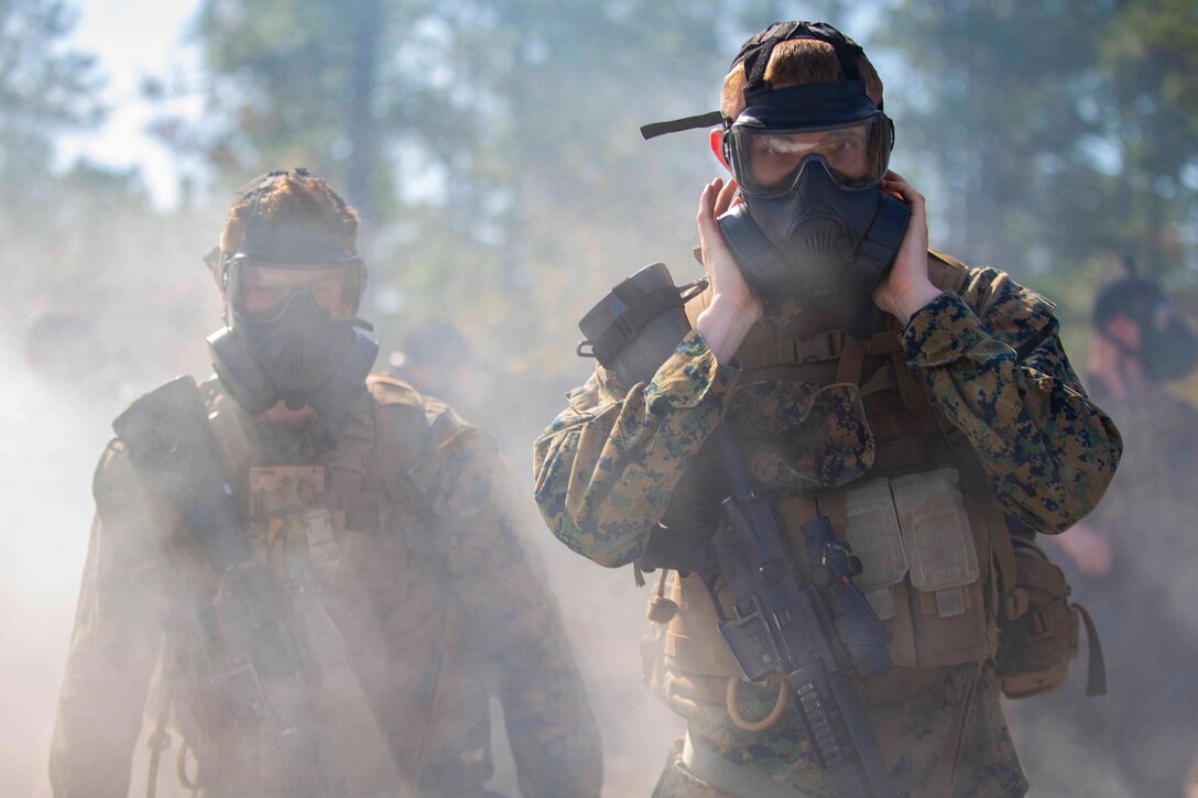 Two Marines wearing gas masks walk through smoke.