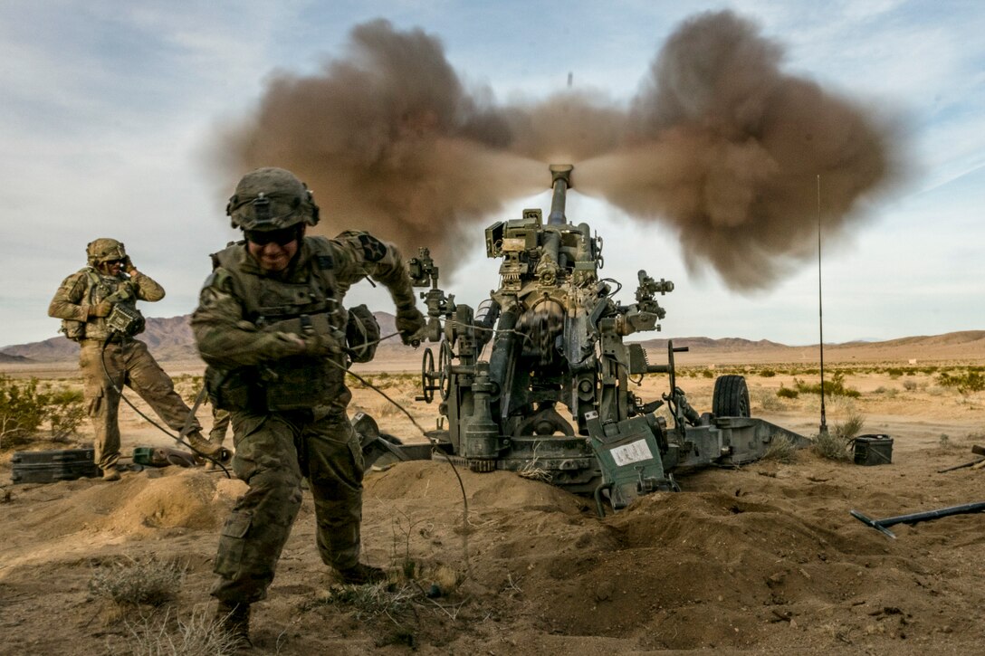 Soldiers brace as a howitzer fires in the desert.